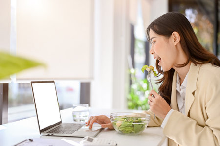 side view of a busy and hungry millennial asian businesswoman eating salad, having lunch at her desk while working on her project on laptop. busy urban lifestyle conceptの写真素材