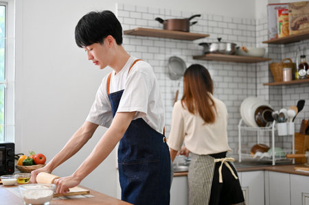 Focused young Asian man in apron is kneading raw dough with a rolling pin, making dough while his...