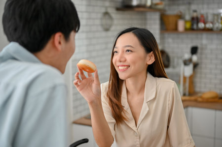 Pretty and lovely young Asian female feeding doughnut to her boyfriend, having happy time together in the kitchen.の写真素材