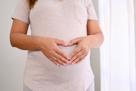 Cropped shot of a pregnant woman in comfy clothes making heart hands sign on her big belly. love,...