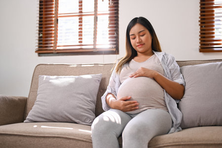 Happy Asian pregnant woman in comfortable clothes relaxes on the sofa in her living room, touching and feeling her baby in her belly with love. motherhood conceptの写真素材