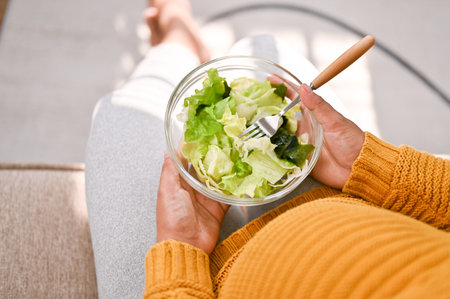 Top view of a pregnant woman holding a salad bowl, eating a healthy salad bowl on sofa in her living room.の写真素材
