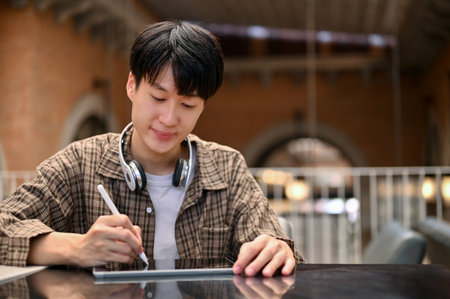Smart and focused young Asian man in casual clothes with wireless headphones using his digital tablet, designing his graphic artwork on tablet, remote working at the coffee shop.の写真素材
