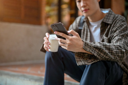 Cropped shot of a handsome young Asian man in casual clothes using his smartphone and sitting on street stairs.の写真素材