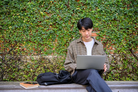 Smart and focused young Asian male college student or freelancer remote working in the city park or garden, using his portable laptop on a bench, working on his tasks.の写真素材