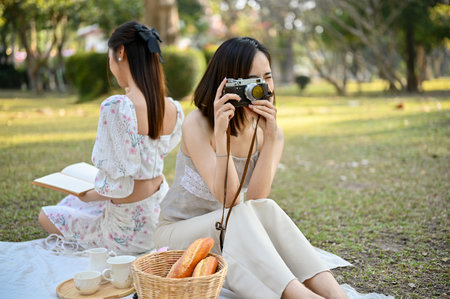 Beautiful hipster Asian woman in casual clothes sits on blanket, enjoying picnic with her friend in the park, taking a picture with her retro camera.の写真素材