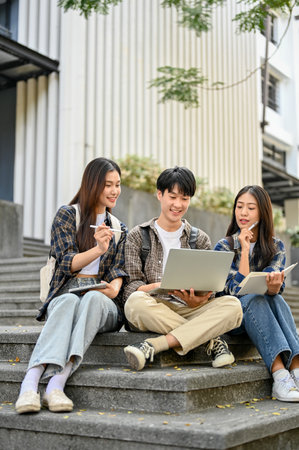 Group of happy young Asian college students sitting on stairs in front of the campus building, talking and focusing on their school project on laptop. university lifestyle conceptの写真素材