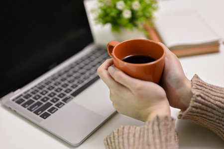 Close-up hands image, A woman in cozy sweater holding a mug of coffee at her working desk.の写真素材