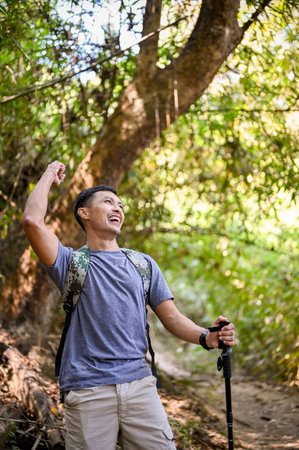 Portrait, Excited and active Asian male traveler or trekker with trekking gear and backpack raising hand, celebrating success, arrived at the destination.の写真素材