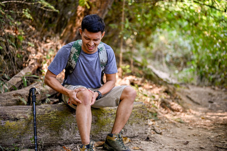 A stressed Asian male trekker sits on a wooden log and holds his knee, suffering from knee pain, injured during trail in the forest. accident, emergency, health problemの写真素材