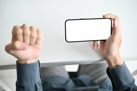 Top view of a man holding a smartphone white screen mockup over the white desk and showing clenched fist hand gesture. victory, winning, success, angry hand gestureの写真素材