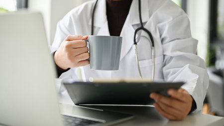 Cropped image of a professional and smart female doctor in uniform sipping coffee and reading medical cases on clipboard at her desk.の写真素材