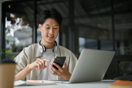 Handsome and focused young Asian male college student or freelancer in casual clothes using his smartphone at the table in the coffee shop.の写真素材