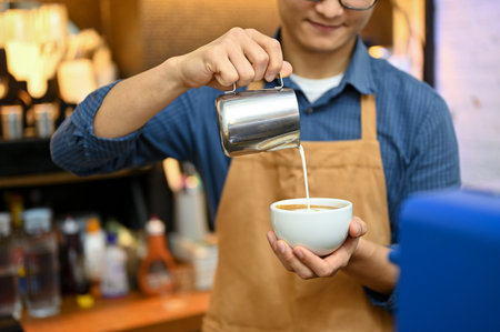 A talented Asian male barista in apron working in the coffee shop, pouring steamed milk into a cup, making latte. cropped imageの写真素材