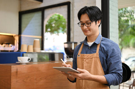 Friendly young Asian male waiter or barista in apron using digital tablet, noting drink orders from...
