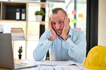 Bored and tired caucasian male engineer or architect sits at his desk, hands on his chin, looking at the camera, feeling no energy to work, overworked, and lacking passion.の写真素材