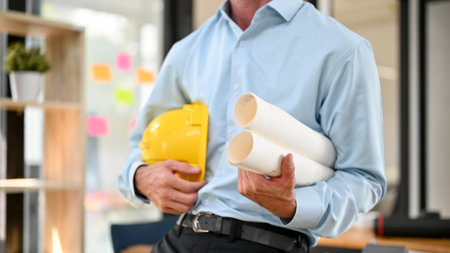 Cropped image of a professional male engineer or architect holding a yellow hardhat and blueprints in the office.の写真素材