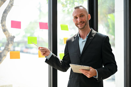 Professional Caucasian male CEO or executive manager in formal business suit is training his employees in the meeting, pointing pen on a sticky note on the glass wall.の写真素材