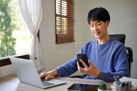 Handsome young Asian male office worker in casual clothes using his smartphone, looking at his phone scree, sitting at his office desk.の写真素材