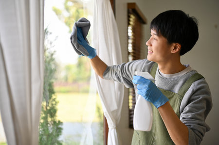 Happy young Asian man wearing rubber gloves using glass spray and dust cloth to clean the window glass, enjoys cleaning house. side viewの写真素材