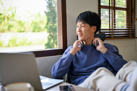 Happy and relaxed young Asian man in comfy clothes with wireless headphones sits at his desk, putting his legs on table, looking through the window and daydreaming about success.の写真素材