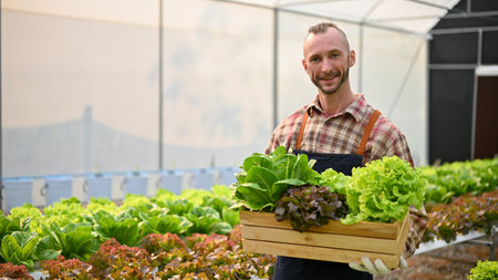 Smiling Caucasian male farm owner or farm worker holding a wooden crate with full of organic fresh...