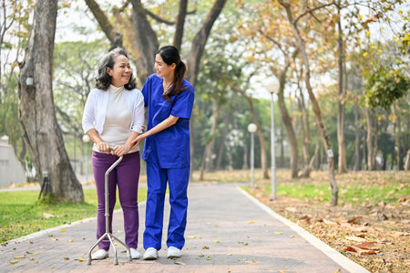 Smiling and caring Asian female caregiver assisting an old lady to walk with a walking stick, strolling around the hospital's park together.の写真素材