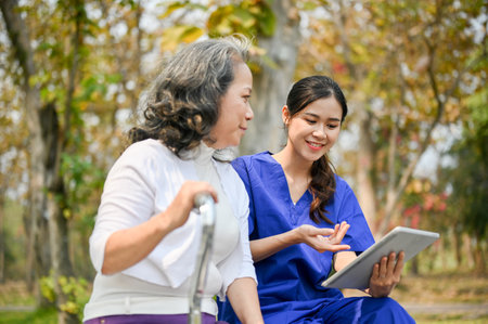 Happy and caring young Asian female nurse or caregiver shows something on her tablet screen to an old lady patient while talking and resting on a bench in the park together.の写真素材