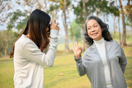 Lovely Asian granddaughter taking a picture of her happy grandmother with retro camera while spending time together in the beautiful green park. leisure conceptの写真素材
