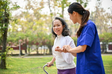 A happy old Asian disabled lady with a walking stick being helped by a caring female caregiver,...