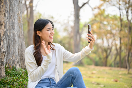 Charming and happy young Asian woman in casual clothes is talking on a video call with her friend through her phone while relaxing in the park.の写真素材