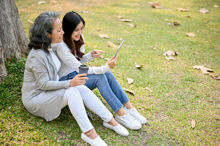 Lovely Asian granddaughter and grandmother sitting under the tree in the green park together, watching video on tablet, sipping coffee and chilling.の写真素材