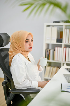 Portrait, Beautiful and successful millennial Asian Muslim businesswoman or female boss wearing hijab sipping her morning coffee at her desk in her private office.の写真素材