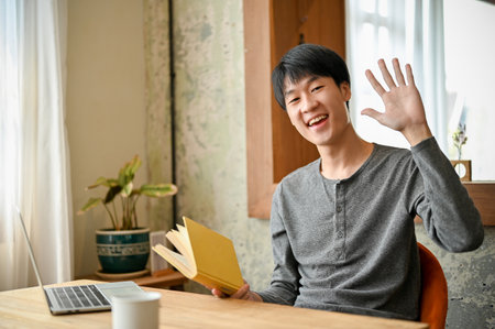 Handsome and cheerful young Asian man in casual clothes sits in the minimal coffee shop with a book, smiling, looking at camera and waving hand.の写真素材