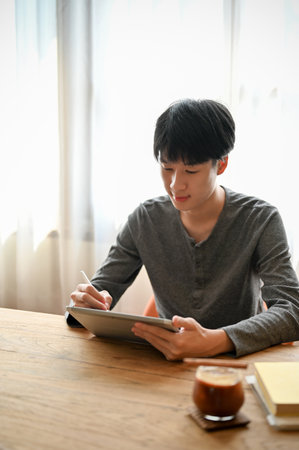 Handsome young Asian man in casual clothes using digital tablet, writing or noting something on his tablet while relaxing in the cafe.の写真素材