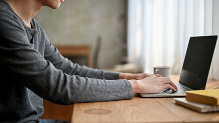 Cropped, side view image of a young Asian man in casual clothes typing on keyboard, using laptop...