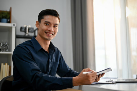 Successful and handsome millennial Asian businessman or male boss holding a digital tablet, sitting at his desk, smiling and looking at the camera.の写真素材