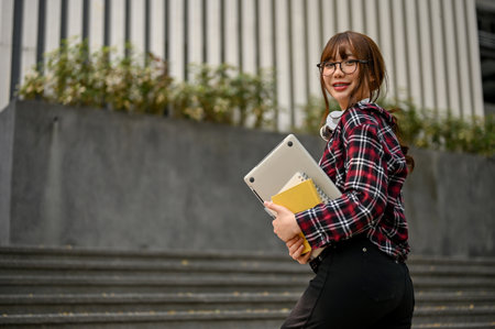 Charming and smiling young Asian female college student walking up the stairs outside the campus building with her belongings.Â uni-lifestyle conceptの写真素材