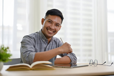 Smart and smiling millennial Asian businessman sits at his desk in his modern office, looking at the camera.の写真素材