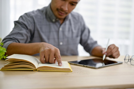 Cropped image of smart and focused millennial Asian businessman using his digital tablet and researching information on a book.の写真素材