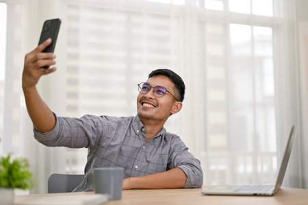 Handsome and smiling millennial Asian man or businessman taking selfie with his phone while sitting at his office desk.の写真素材