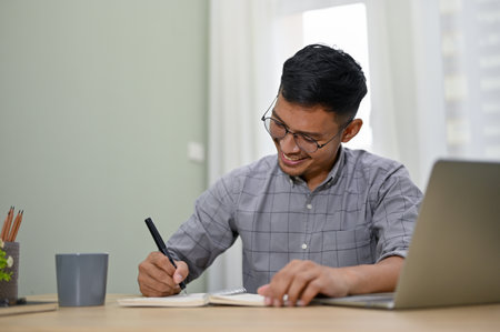Happy and professional millennial Asian businessman or male office worker writing something on his book, working at his desk in the office.の写真素材