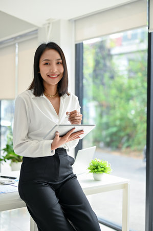 Portrait, Beautiful and confident millennial Asian businesswoman in trendy clothes sits on table with her tablet, smiling and looking at the camera.の写真素材