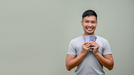 Smiling and happy millennial Asian man in casual clothes holding cash, USD dollar bills while standing against a green studio background. wealth, investment, profit, incomeの写真素材