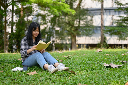 Pretty young Asian female college student in casual clothes sits on grass and reads a book in the campus park.の写真素材