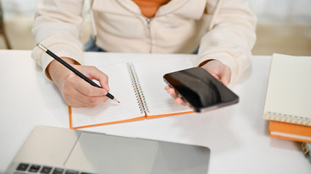 Top view of a woman in casual clothe using her phone and making list on her notepad, working at her...