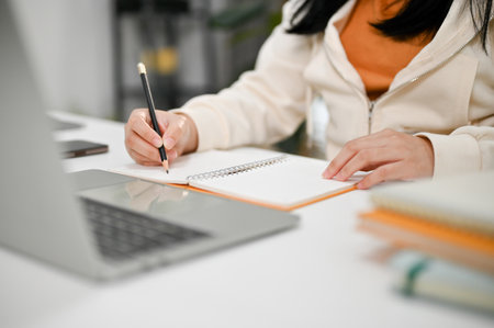 Cropped image of an Asian female college student doing homework, writing something on her notebook and using laptop computer.の写真素材