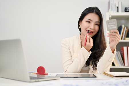 Attractive and charming millennial Asian businesswoman or female secretary looking at the mirror, using lipstick, applying makeup on her face at her desk in the office.の写真素材