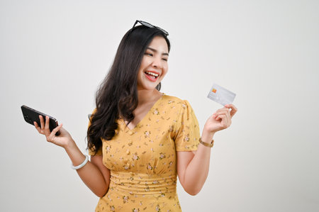 Happy and smiling young Asian female holding a credit card and smartphone while standing against an...