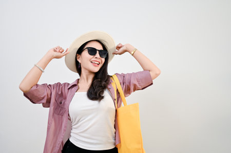 Attractive and cheerful young Asian female tourist in sunglasses touching her straw hat and carrying her tote bag while standing against an isolated white background. shopper, traveler conceptの写真素材